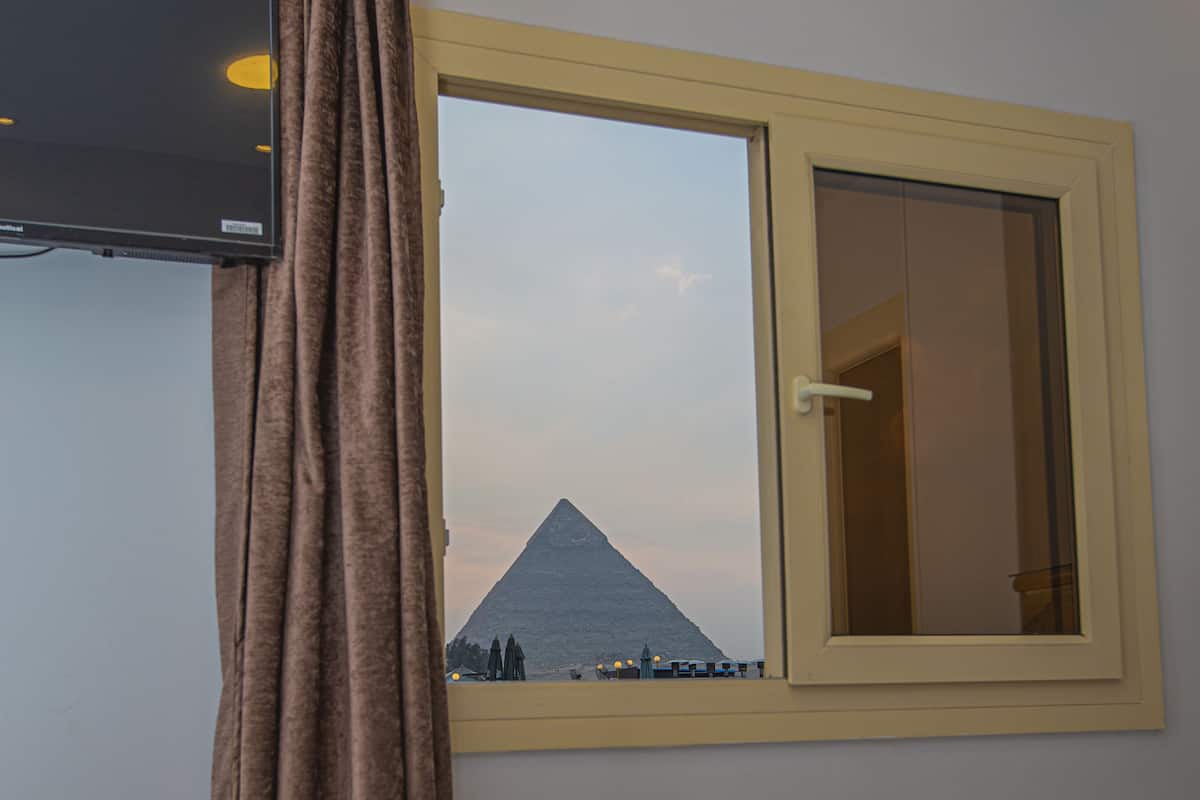Hotellihuone, jossa on sänky, ikkuna, josta avautuu näkymä pyramidiin, ja verho.