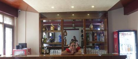 A bar area with shelves stocked with various bottles and glasses, a Coca-Cola refrigerator, and a counter where a person is leaning.