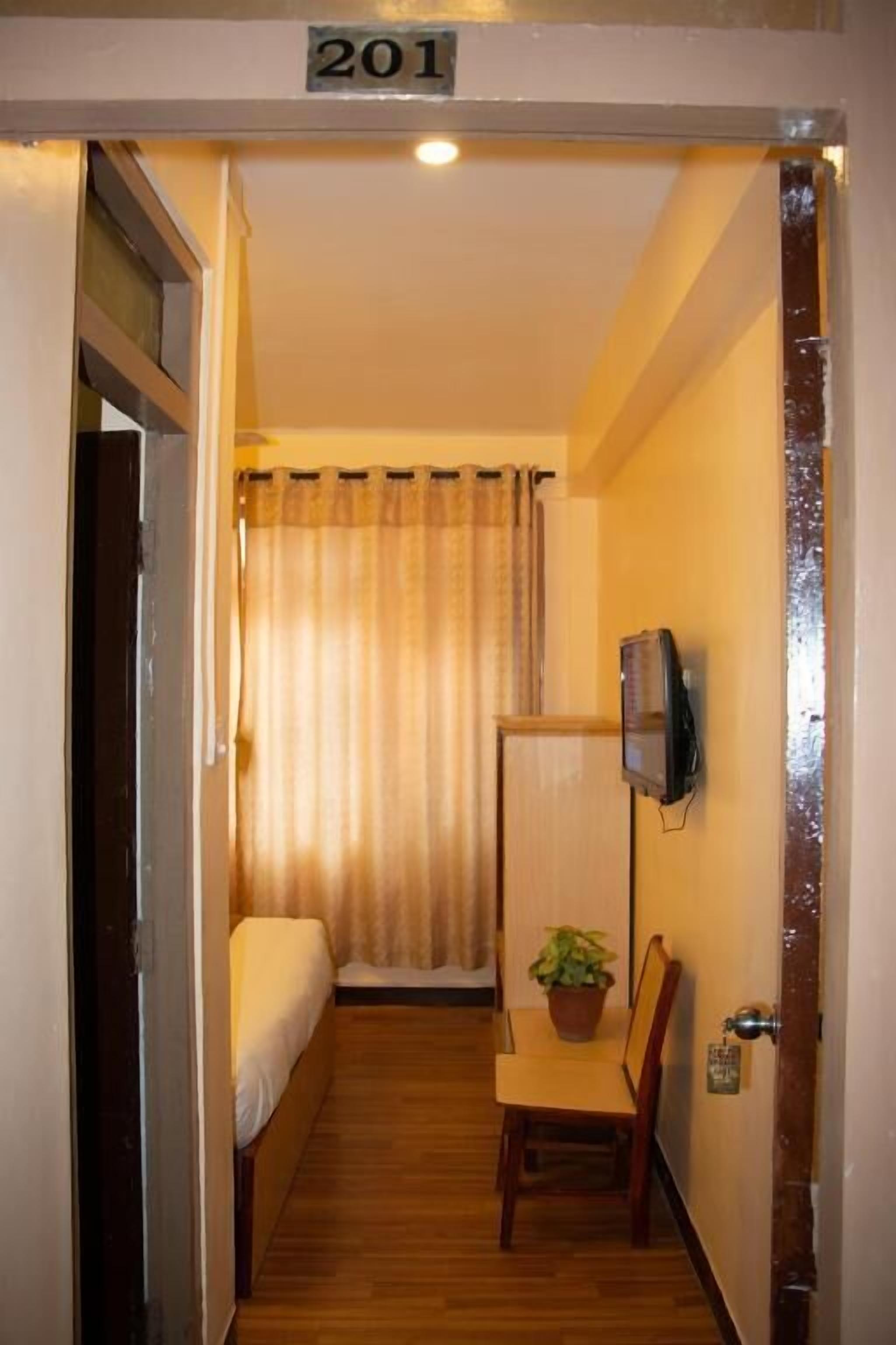 A small, narrow room with a bed, a television mounted on the wall, and a wooden chair with a plant.