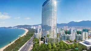 Un edificio moderno de gran altura con fachada curva, situado cerca de la costa, con una playa y montañas al fondo.