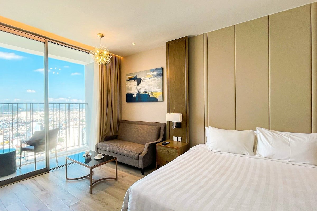 Habitación de hotel con una cama grande, una silla, una mesita, un cuadro y vistas a un paisaje urbano a través de un ventanal.