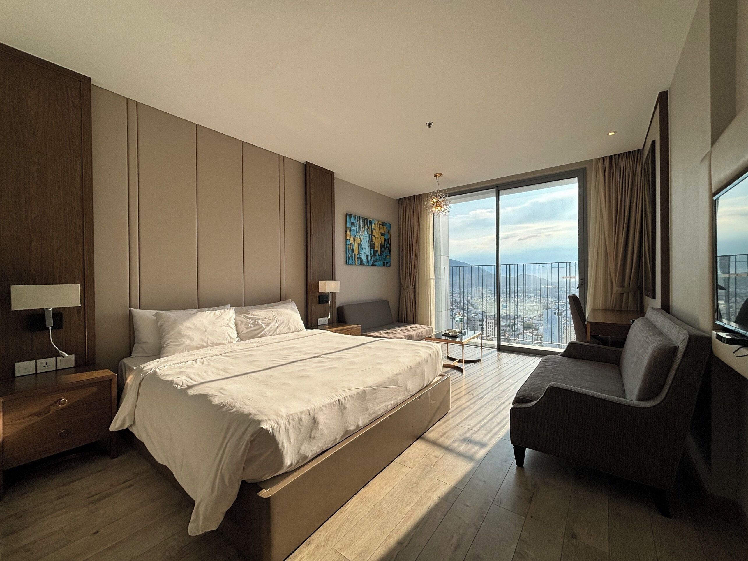 A modern hotel room with a large bed, a seating area, and a city view through large windows.