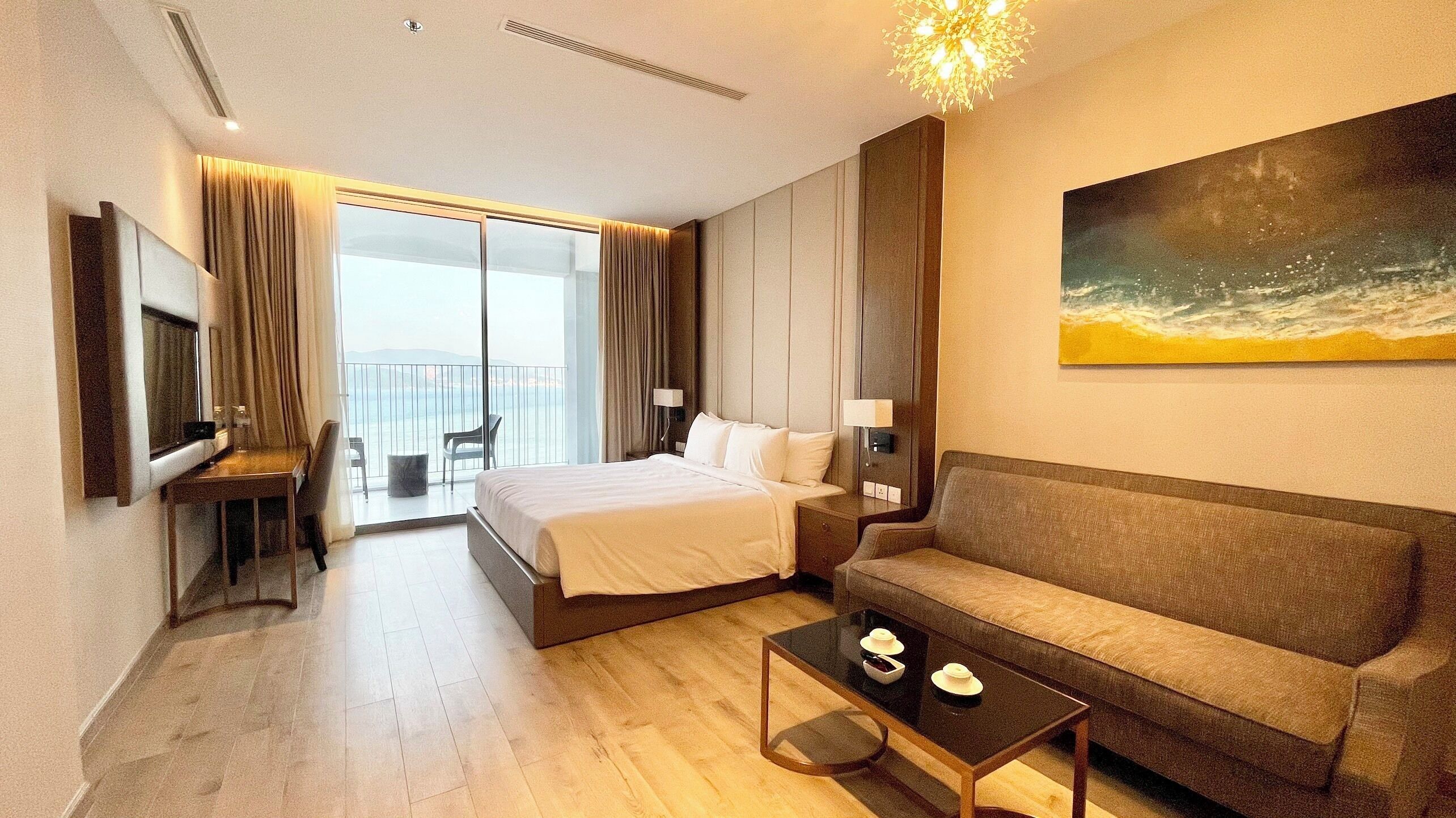 A hotel room with a bed, a sofa, a TV, a painting, and a balcony with a view.
