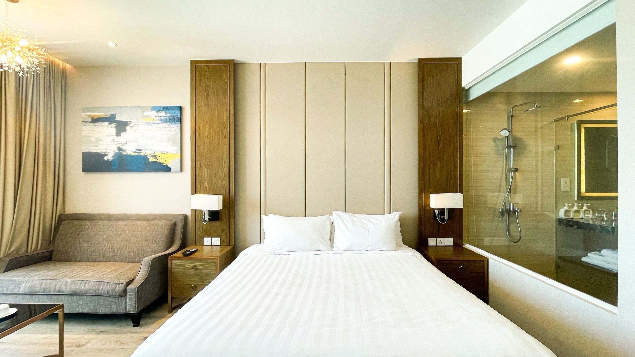 A hotel room with a bed, a chair, a nightstand, a lamp, a painting, and a shower area.