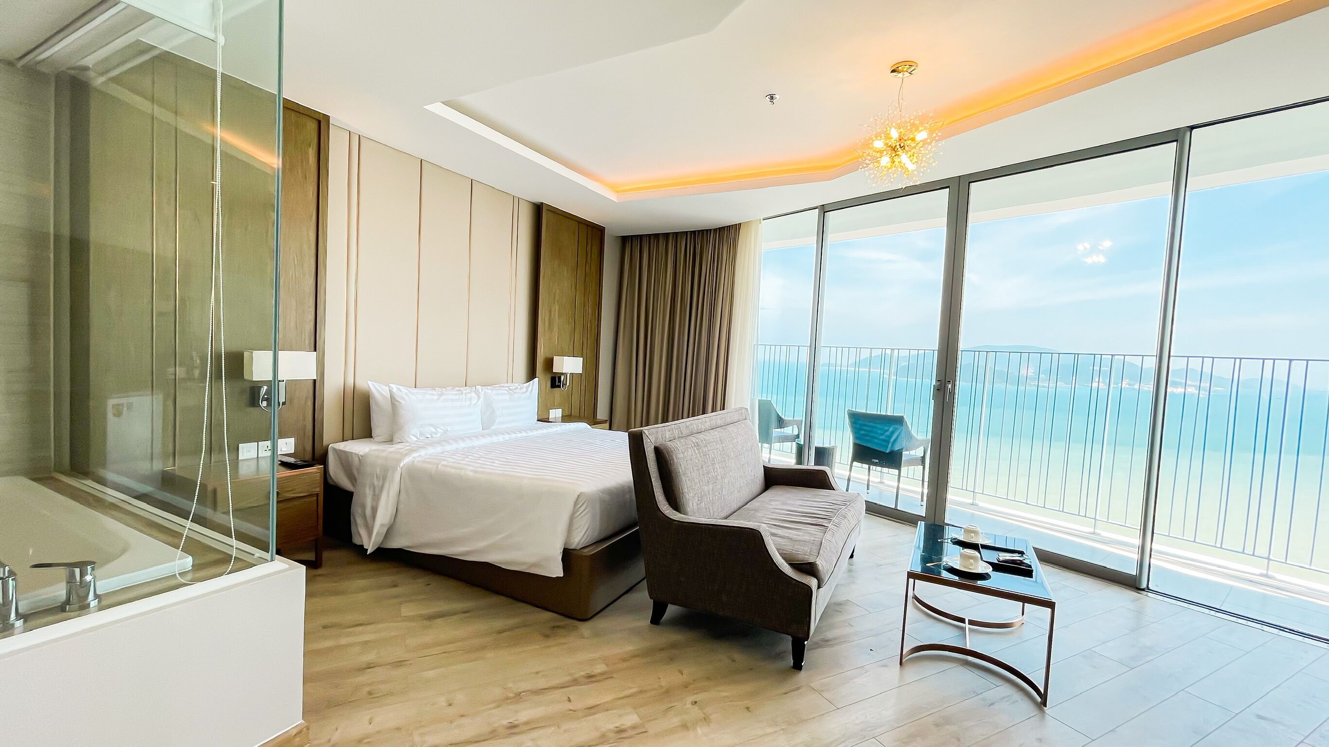Una habitación de hotel con cama, sofá, separador de vidrio, balcón con vistas y una lámpara de araña moderna.