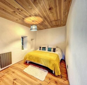 Une chambre confortable avec un plafond en bois, un lit recouvert d’une couverture jaune, une petite fenêtre et un radiateur.