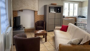 Una sala de estar con sofá, sillón y mesa de centro. Hay un televisor, chimenea y una zona de cocina con ventana.