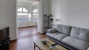 Ein Wohnzimmer mit grauem Sofa, einem Couchtisch und einem Fernseher. Ein Fenster bietet Blick auf das Meer.