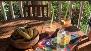 A wooden table with a bowl of fruit, a tray of fried snacks, a glass of drink, and a bottle of alcohol on a balcony with a view of the ocean.