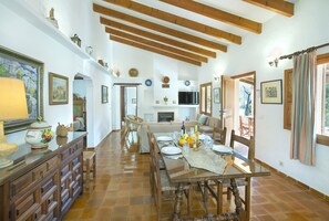Amplia sala de estar con una mesa de comedor dispuesta para cuatro personas, un sofá y un televisor. La habitación tiene vigas de madera en el techo y suelo de baldosas.