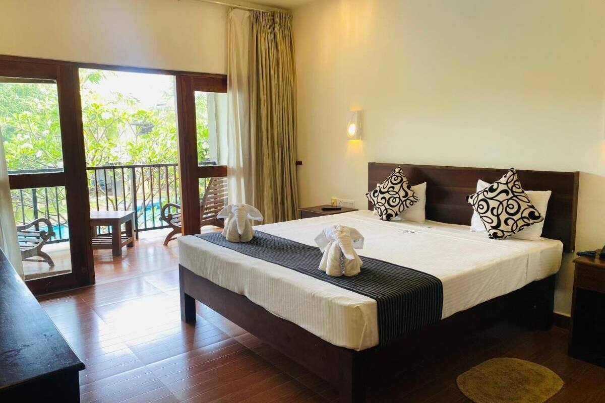 A hotel room with a large bed, two pillows, a bedside table, a nightstand, and a view of the outdoors through a sliding door.