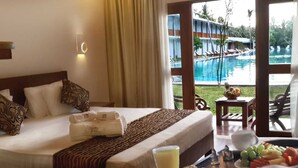 A hotel room with a bed, a breakfast tray with fruits and a glass of juice, and a view of a swimming pool outside.