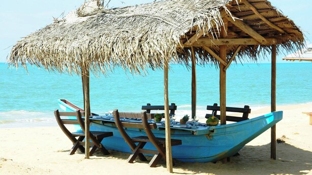 A thatched-roof structure with a blue boat and wooden benches on a sandy beach.
