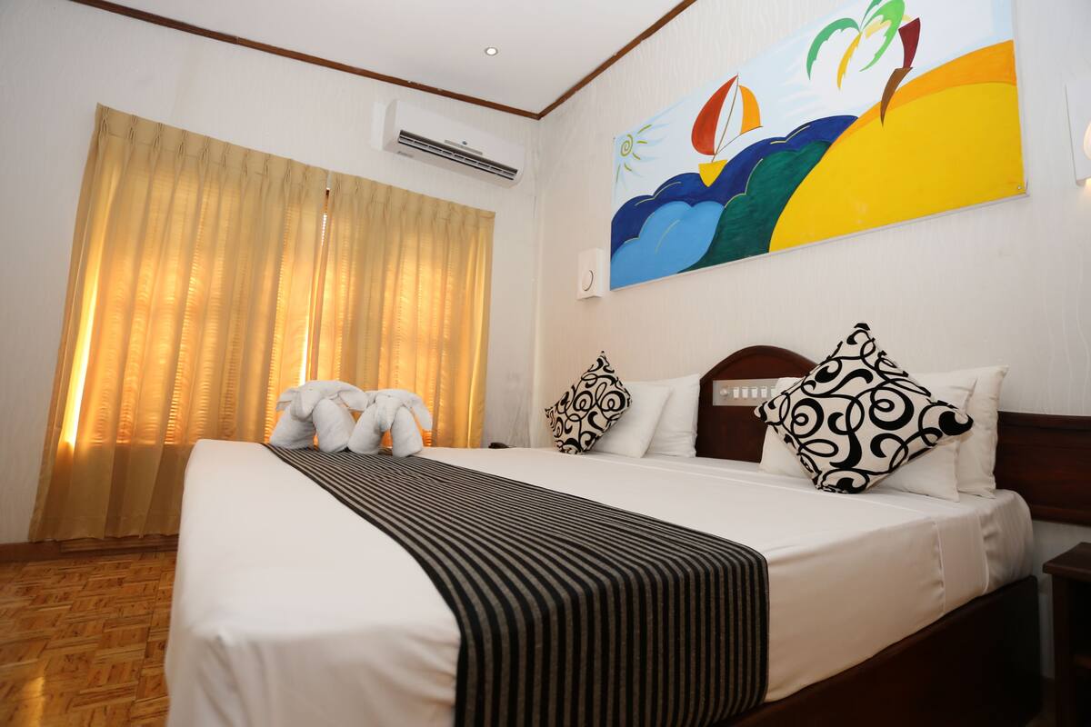 A hotel room with a large bed, a wooden headboard, a painting on the wall, and a window with curtains.