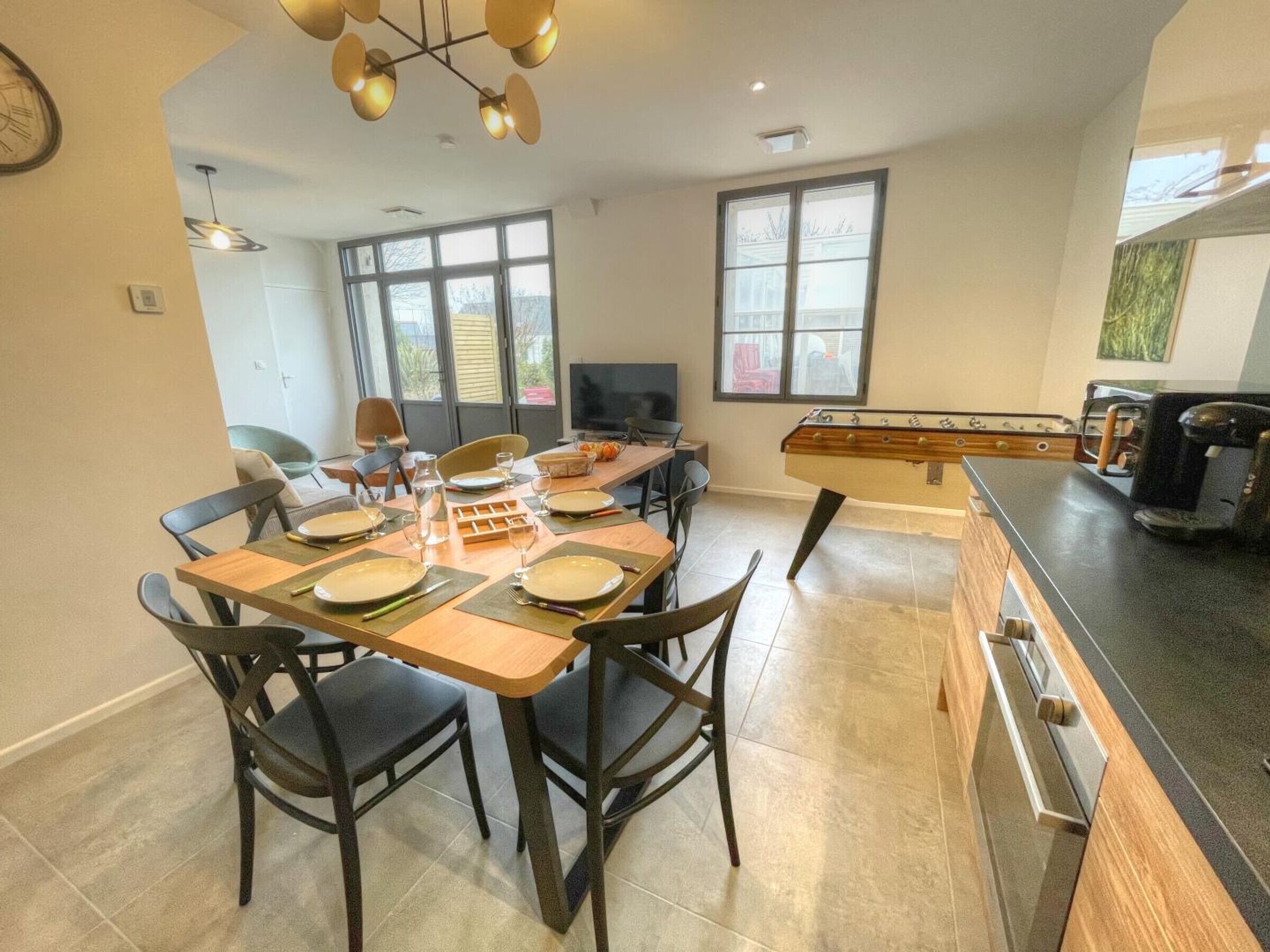 Un coin repas avec une table en bois dressée pour quatre personnes, entourée de chaises. Un baby-foot se trouve à droite, et un espace cuisine comprend une machine à café.