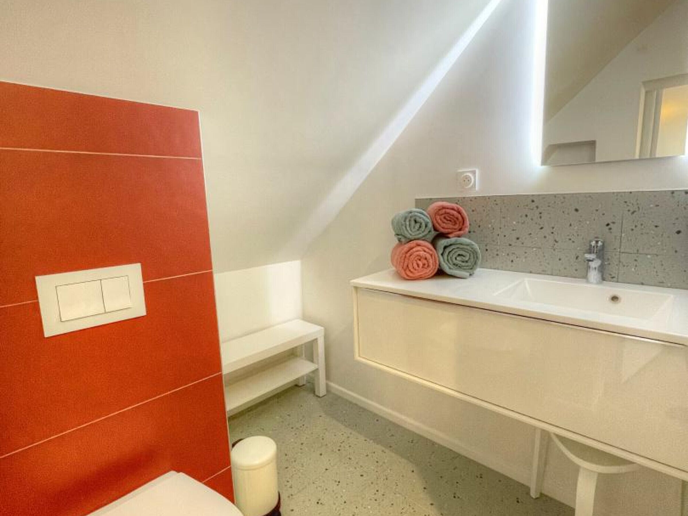 A bathroom with a white sink, a mirror, and a bench. There are towels stacked on the sink.