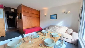 A compact living space with a dining table set for four, a sofa, and a wooden cabinet with a red cushion. - Spacious 6-Person Studio with Bunk Beds and Ski Locker, Dishwasher, and Fondue Set (Piau-Engaly)