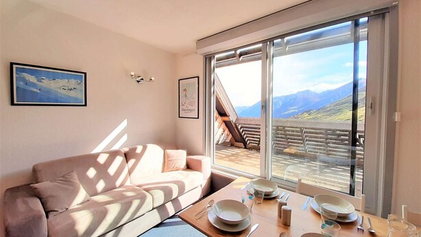 A modern living room with a sofa, dining table set for two, and a large sliding glass door leading to a balcony with a mountain view. - Spacious 6-Person Studio with Bunk Beds and Ski Locker, Dishwasher, and Fondue Set (Piau-Engaly)