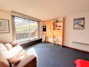 Ein Zimmer mit einem großen Fenster, einem Holzkasten, einem weißen Hocker und einem roten Stuhl.