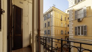 A narrow balcony with a metal railing, adjacent buildings with multiple windows, and a door with a shutter.