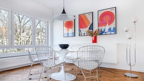 A modern dining area with a white table, wireframe chairs, and a hanging light fixture. There are three framed artworks on the wall and a vase with flowers on the table. - Molière Design residence  (Brussels)