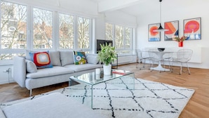 A modern living room with a grey sofa, colorful cushions, a glass coffee table, a white rug, and a dining area with white chairs and a table. - Molière Design residence  (Brussels)