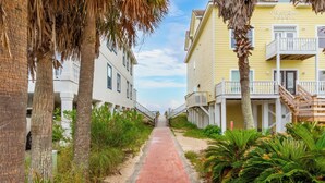 A coastal walkway leading to a yellow building with a balcony, surrounded by palm trees and greenery. - DUNES 102, 1/2 1st Floor, No Elevator, Sea South Properties (Gulf Shores)