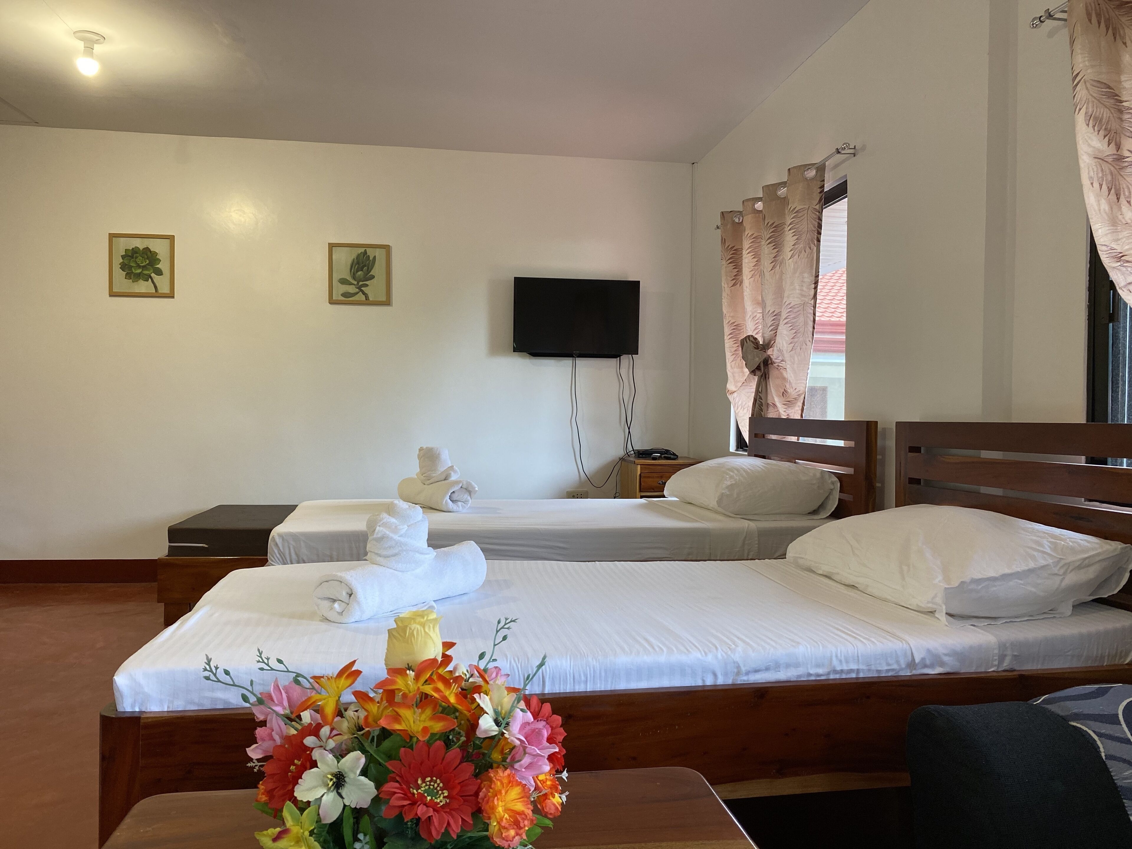 A hotel room with two beds, a flat-screen TV mounted on the wall, a bedside table with a lamp, and a window with curtains.