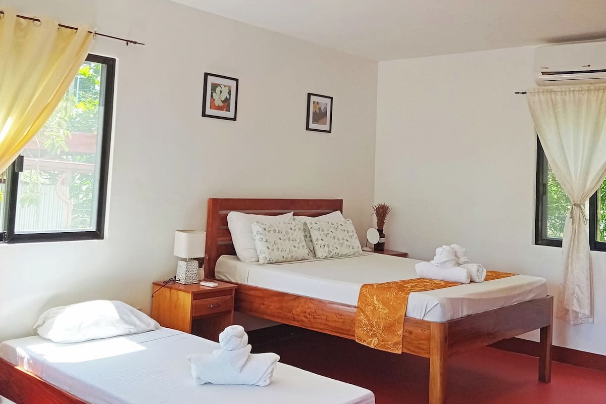 Une chambre d’hôtel avec deux lits, une tête de lit en bois, une table de chevet avec une lampe, une fenêtre avec des rideaux et un appareil de climatisation.