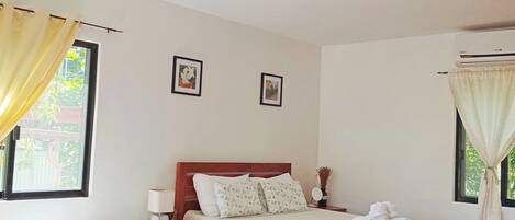 A hotel room with two beds, a wooden headboard, a bedside table with a lamp, a window with curtains, and an air conditioning unit.