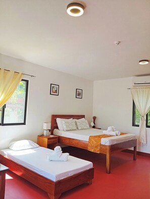 A hotel room with two beds, a wooden headboard, a bedside table with a lamp, a window with curtains, and an air conditioning unit.