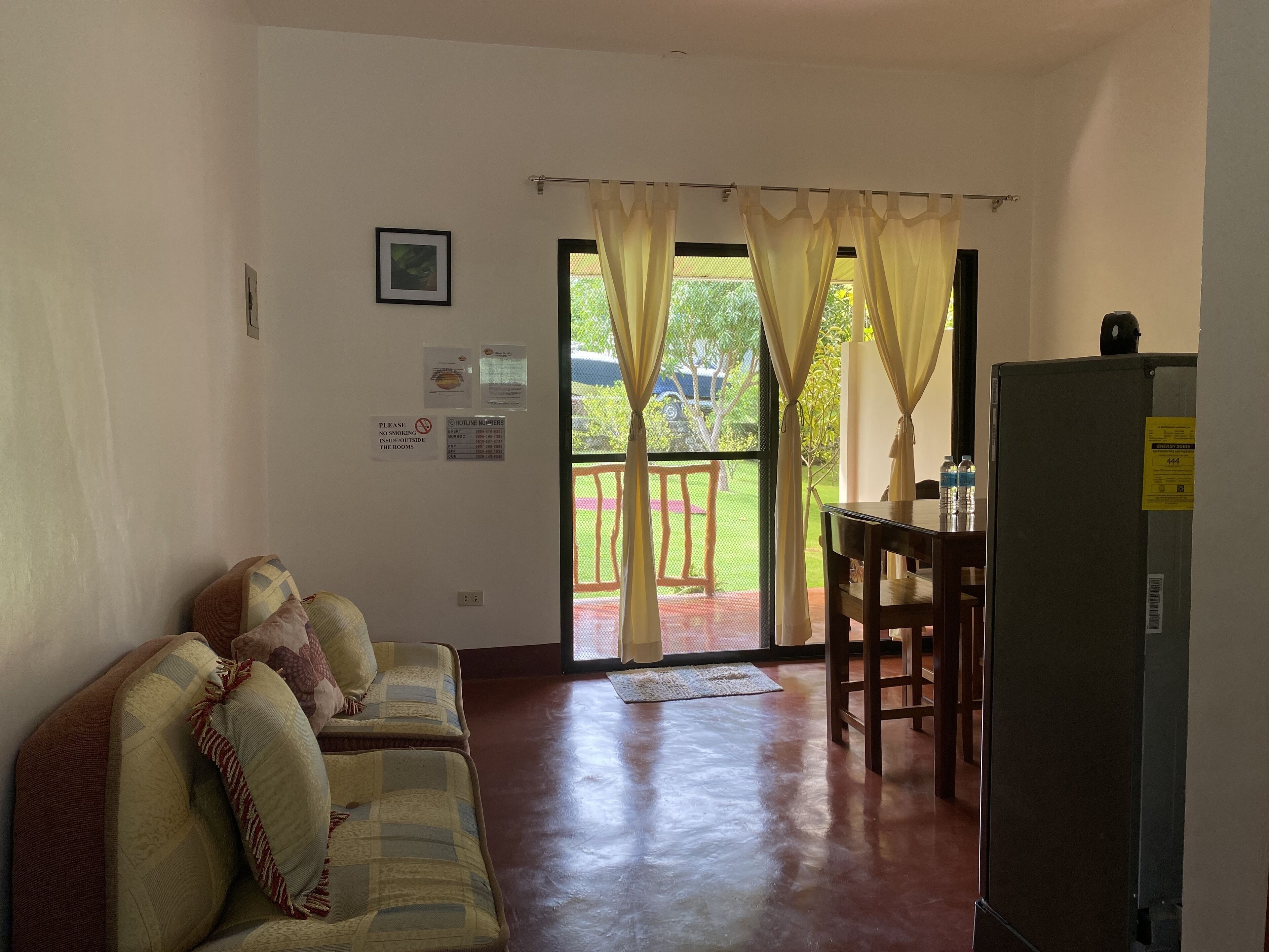 A living room with a sofa, a table, and a refrigerator. There are curtains on the door, and a window with a view of greenery outside.