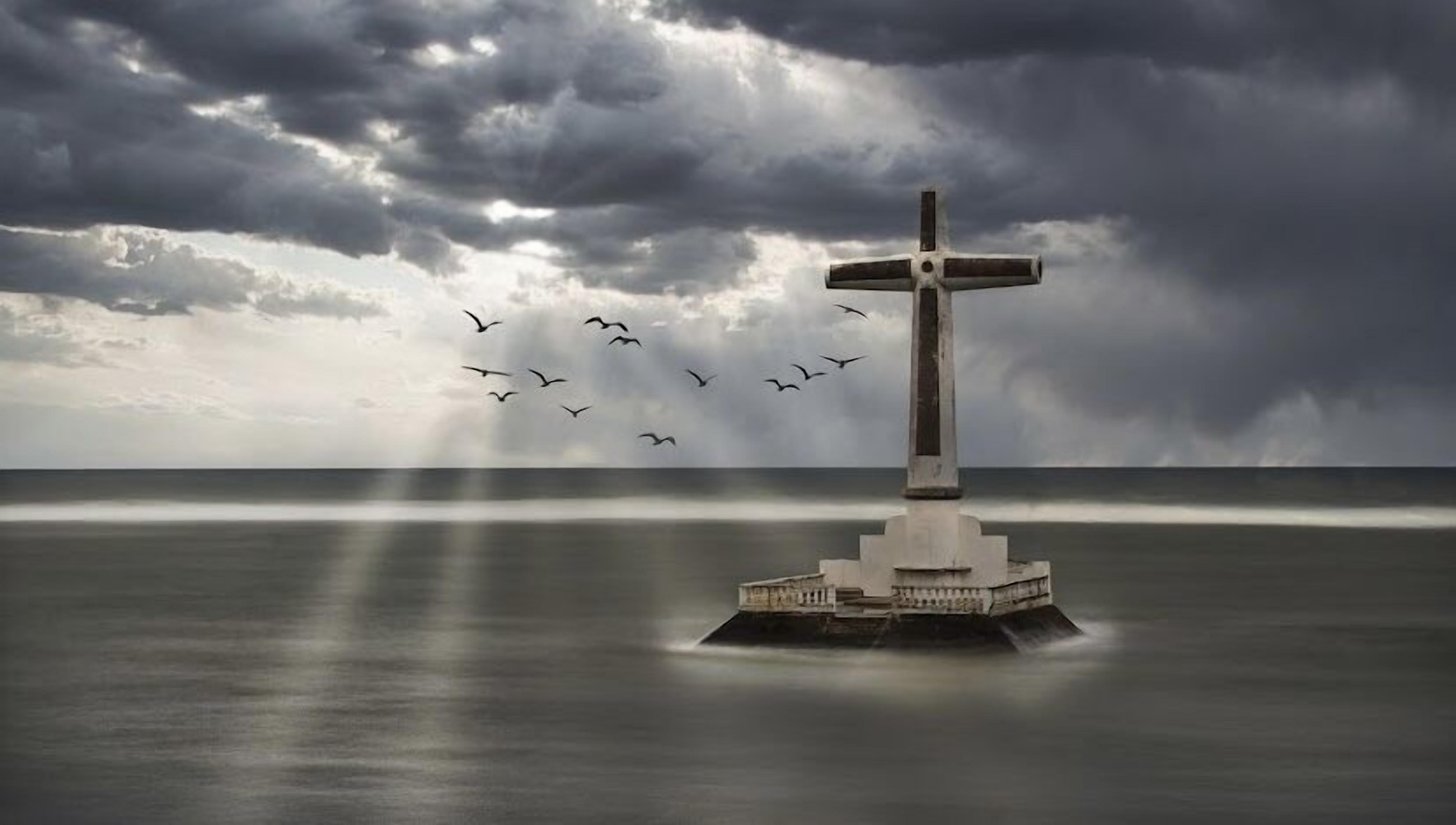 Un monumento en forma de cruz sobre una plataforma en medio de un cuerpo de agua bajo un cielo nublado.