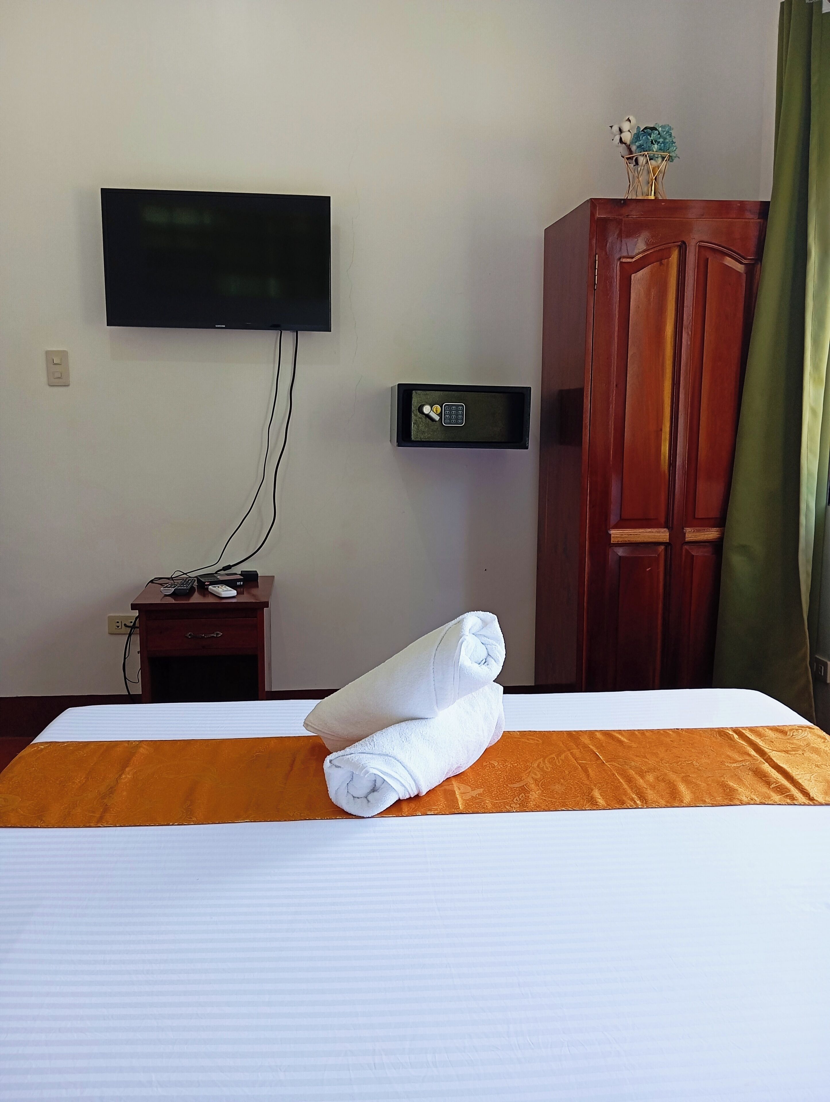Une chambre d’hôtel équipée d’un lit, d’un téléviseur fixé au mur, d’un coffre-fort, d’une armoire en bois et d’un bureau avec une télécommande.