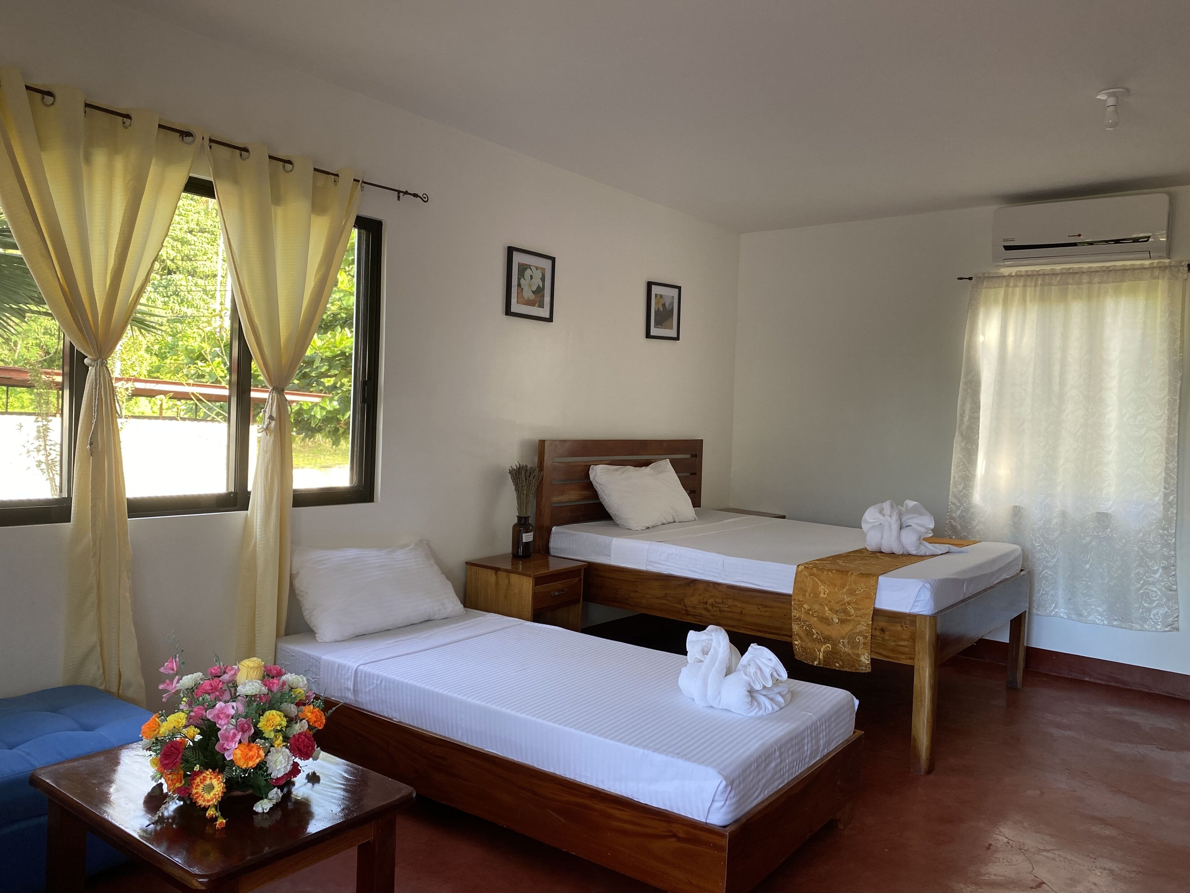 A hotel room with two single beds, a wooden nightstand, a small table with a vase of flowers, and an air conditioning unit.