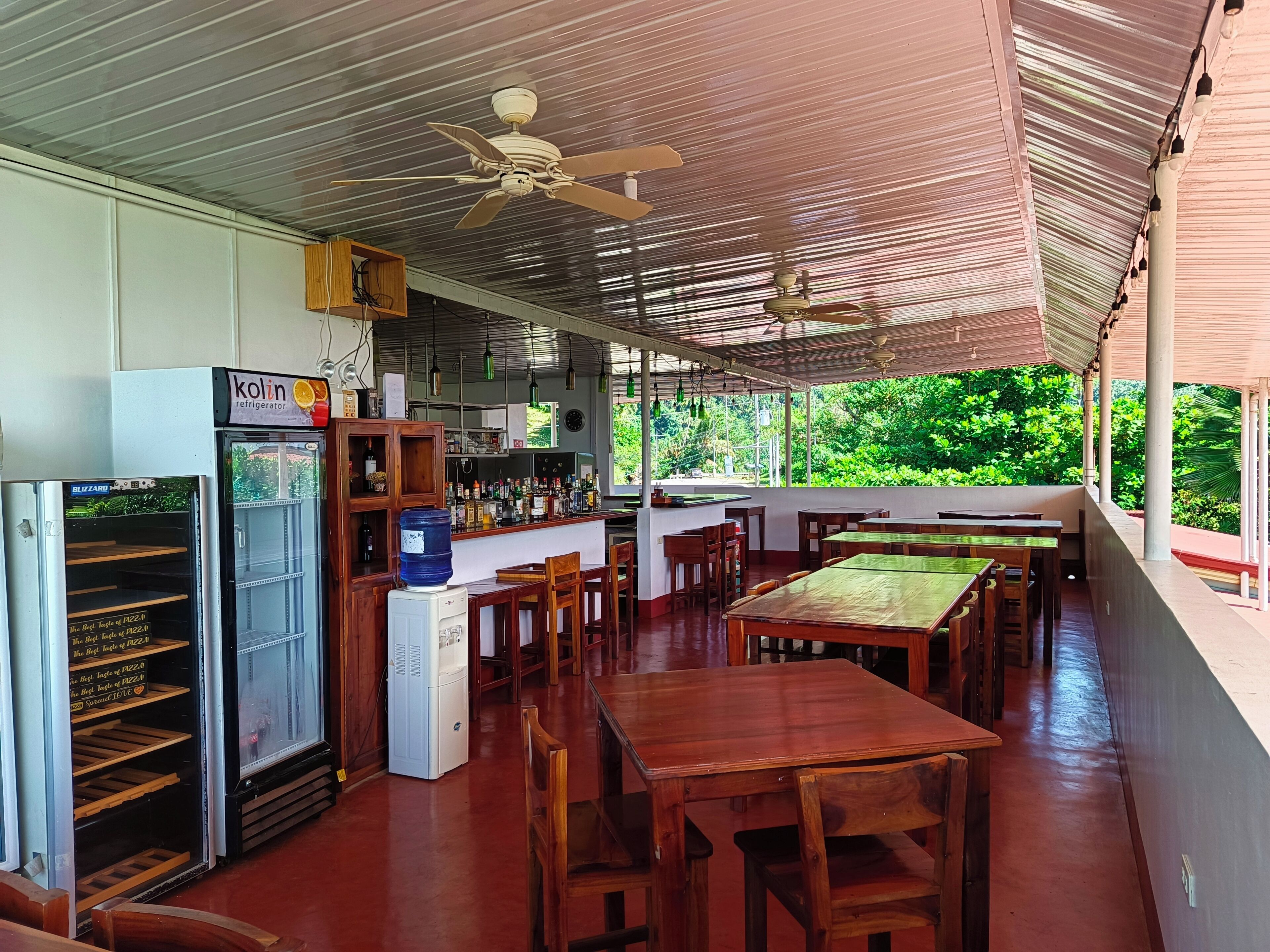 Un espace extérieur abrité, aménagé pour les repas, avec des tables et des chaises en bois, un réfrigérateur et un coin bar doté d’étagères remplies de bouteilles.