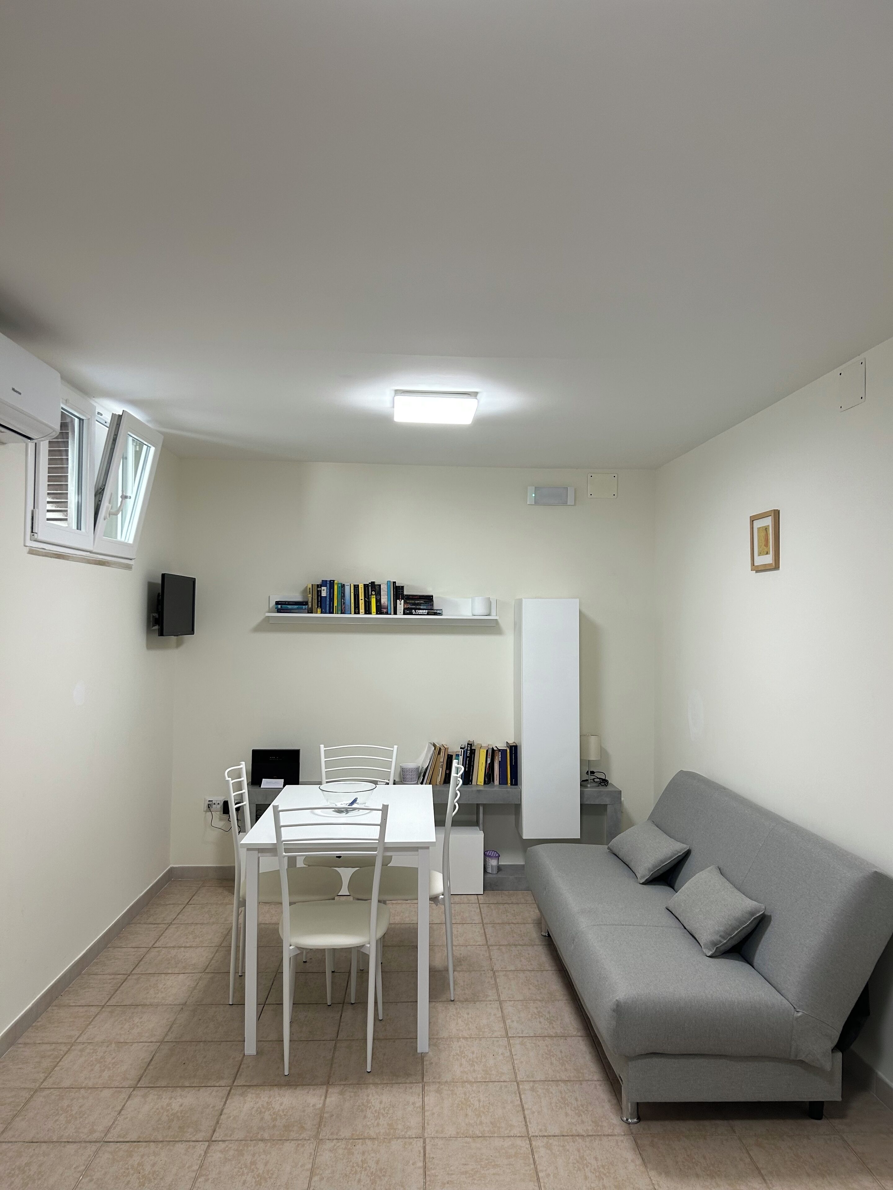 Un espace de vie compact comprenant une table et des chaises, un canapé gris, une bibliothèque et un téléviseur à écran plat.