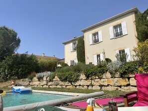A pool area with a stone wall, a large house with white shutters, and a clear blue sky.