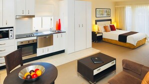 Une chambre d’hôtel avec une kitchenette, un lit doté d’une tête de lit, une table de chevet, une commode et une fenêtre avec des rideaux.