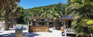 Un petit kiosque situé au bord de la plage, doté d’un toit de chaume, entouré de palmiers et bordé d’une zone sablonneuse où sont disposées des chaises de plage.