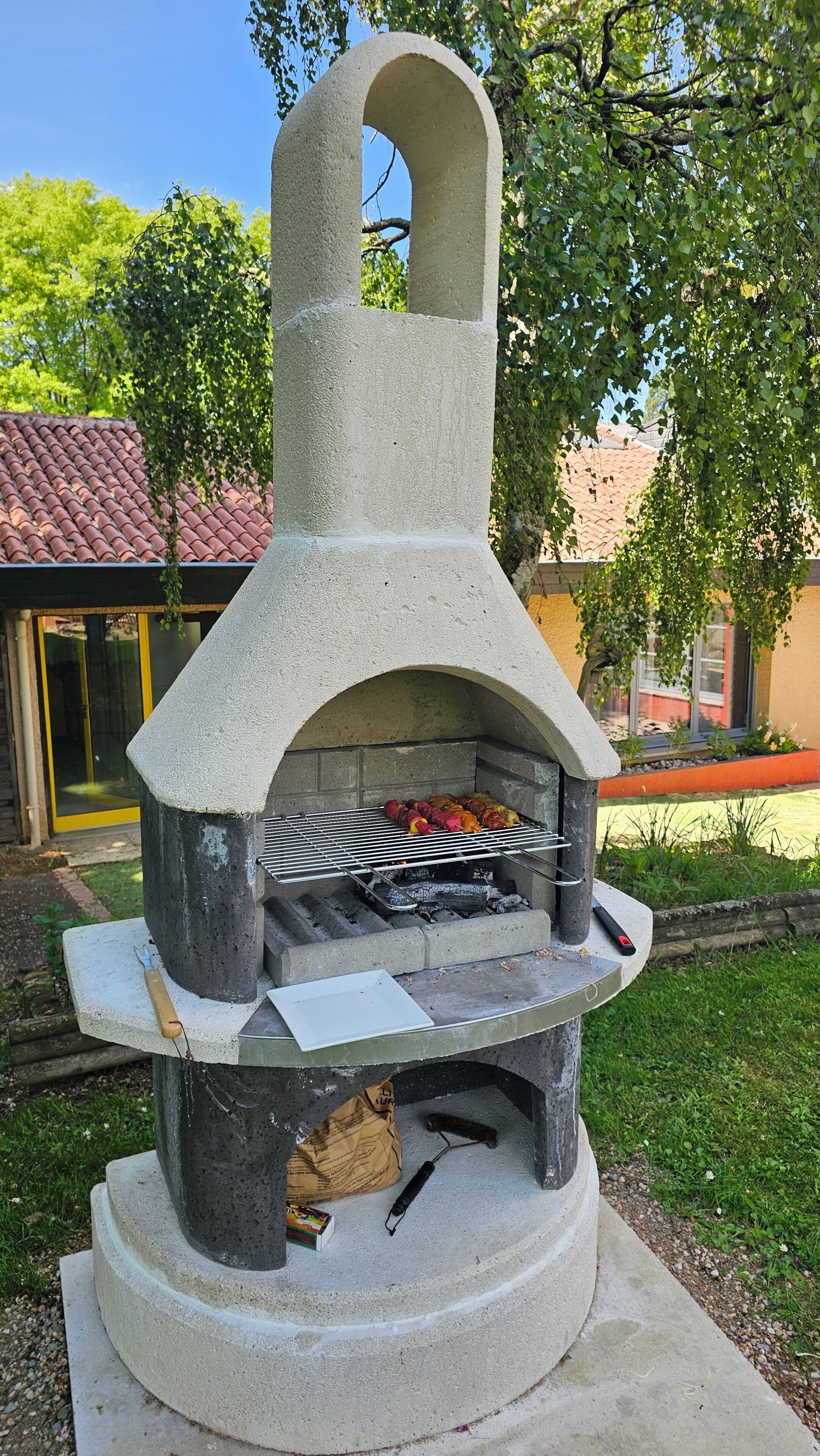 Un four à pizza extérieur en pierre, doté d’une grille en métal, entouré de verdure et avec, en arrière-plan, un bâtiment dont le toit est recouvert de tuiles rouges.