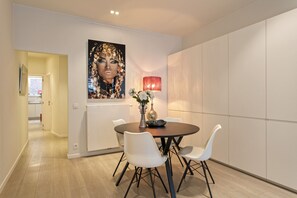 A modern dining area with a round table, white chairs, a vase with flowers, and a large framed artwork on the wall. - Museum Zuid Top located apartments (Antwerp)