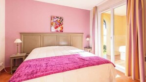 Ein Schlafzimmer mit einem Bett, einem Nachttisch, einer Lampe, einem farbenfrohen Kunstwerk an der Wand und einer Glastür, die zu einem Außenbereich führt.