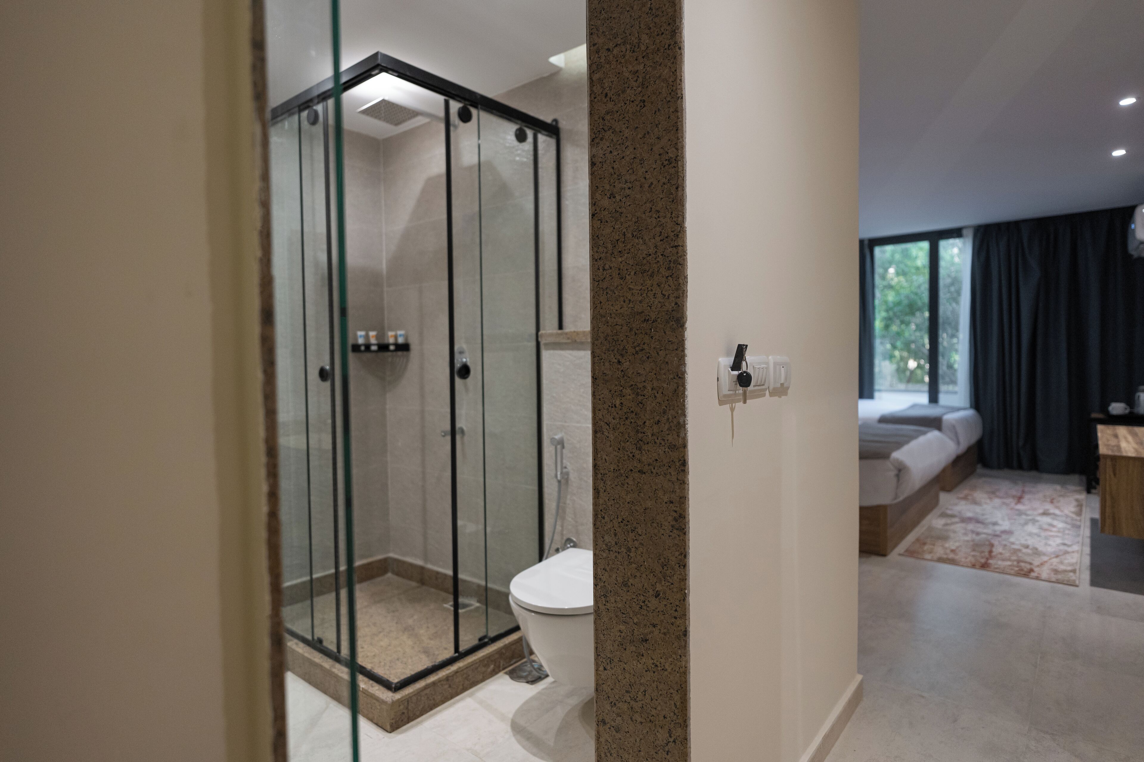 A modern bathroom with a glass-enclosed shower, a toilet, and a bed in the adjacent room.