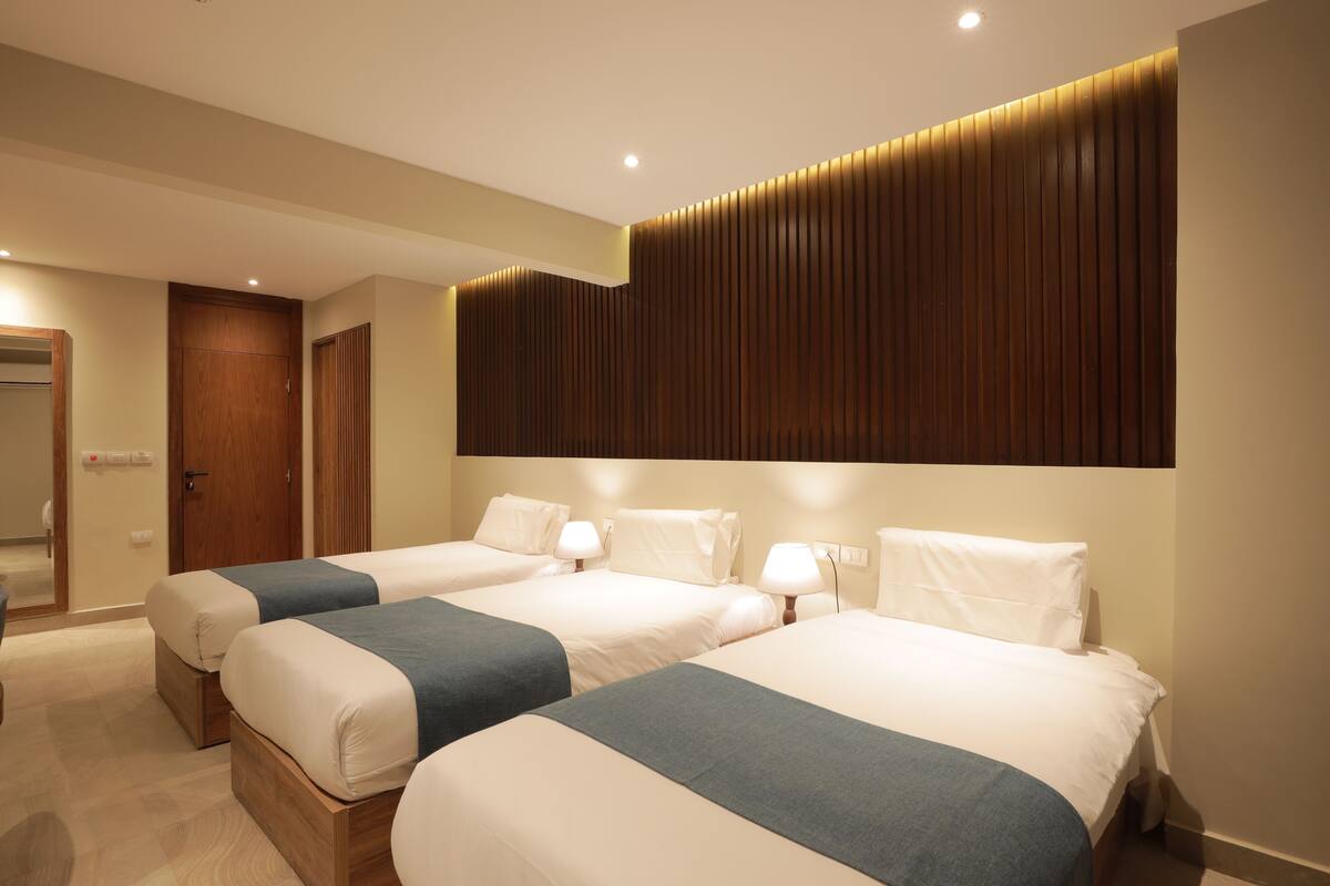 Quarto de hotel com três camas dispostas em linha, cabeceira de madeira e porta à esquerda.