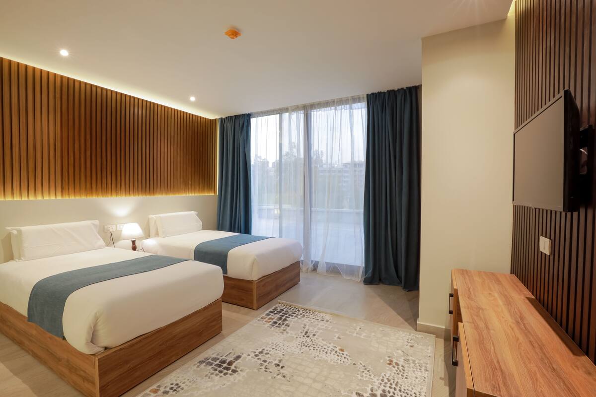 Quarto de hotel moderno com duas camas, cabeceira de madeira, televisão e varanda com cortinas.