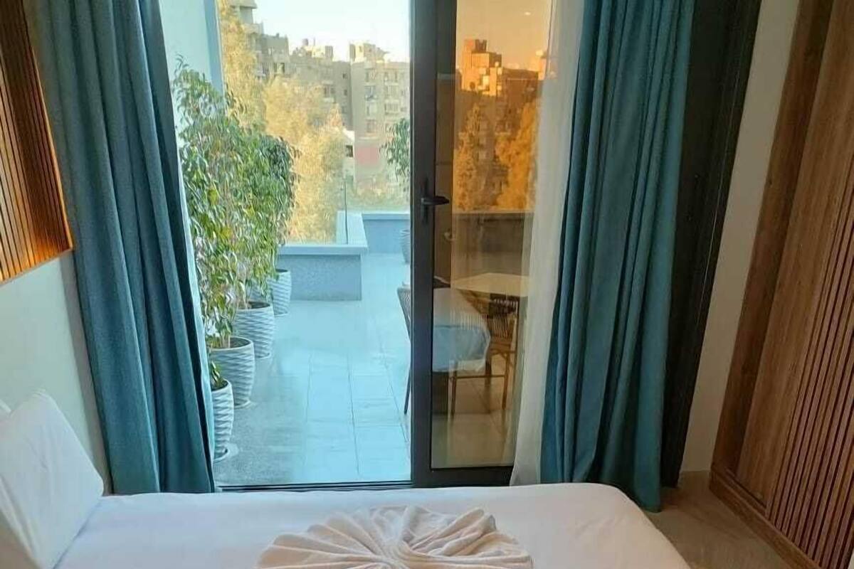 Quarto de hotel com uma cama, vista da cidade pela janela e uma varanda com plantas em vasos.