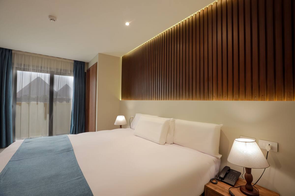 Quarto de hotel com uma cama grande, duas luminárias de cabeceira, um telefone e vista para montanhas através de cortinas finas.