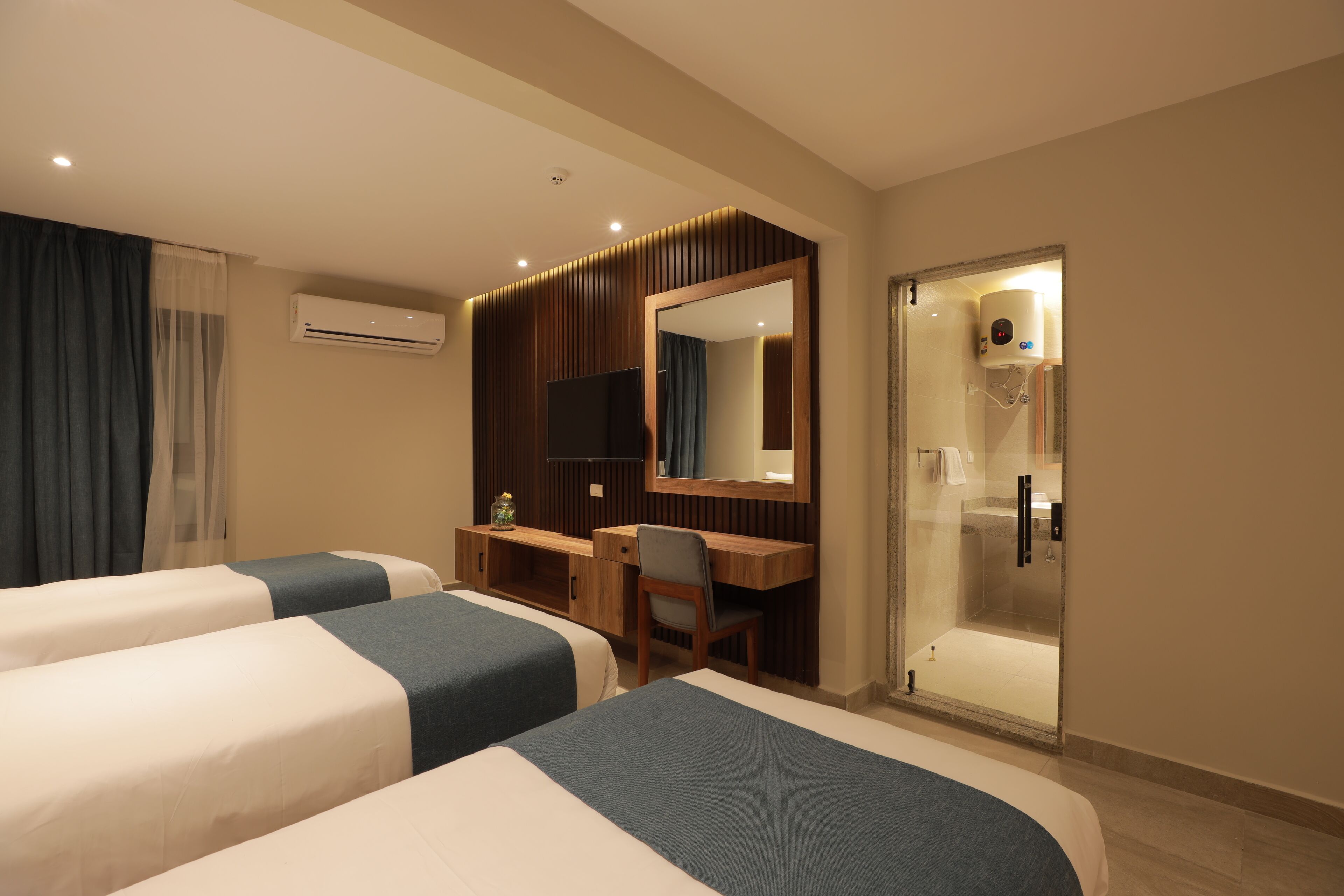 Habitación de hotel con dos camas, un escritorio, una silla, un televisor, un espejo y cabina de ducha con paredes de vidrio.
