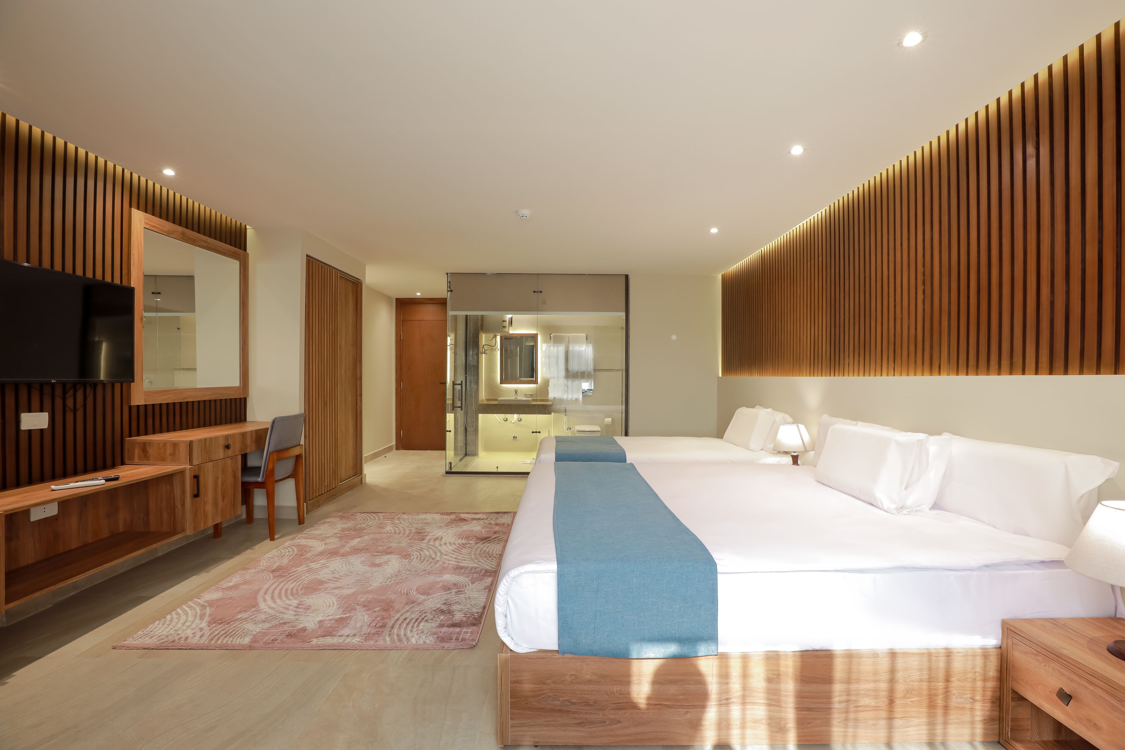 A modern hotel room with a large bed, wooden paneling, a desk with a chair, a TV, and a bathroom visible through a glass door.